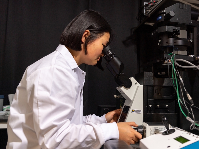 woman in a laboratory is leaned over while operating a microscope 