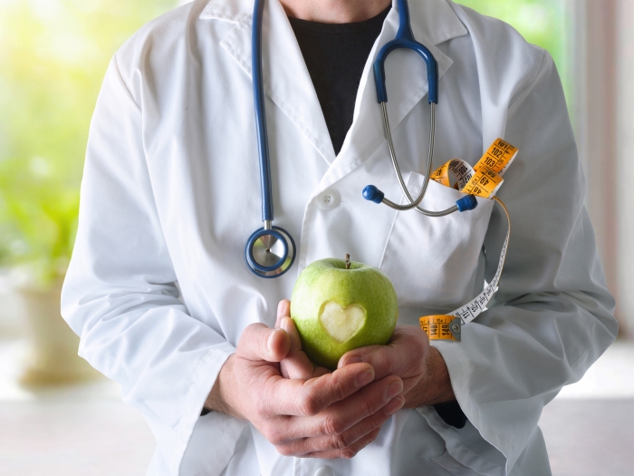 apple in hand of doctor with white coat and stethoscope facing frontward, neck down, and green plant with sunlight behind it from window coming through on white windows sill