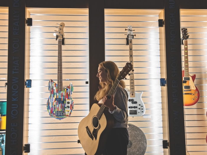 Erica Ginepro stands in front of a wall display of different types of guitars. She's holding an acoustic guitar, is facing sideways, and is looking to the side.