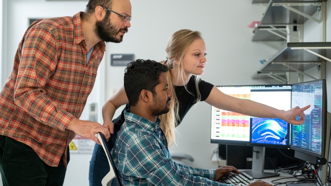 A student points to the computer while discussing research with 2 others