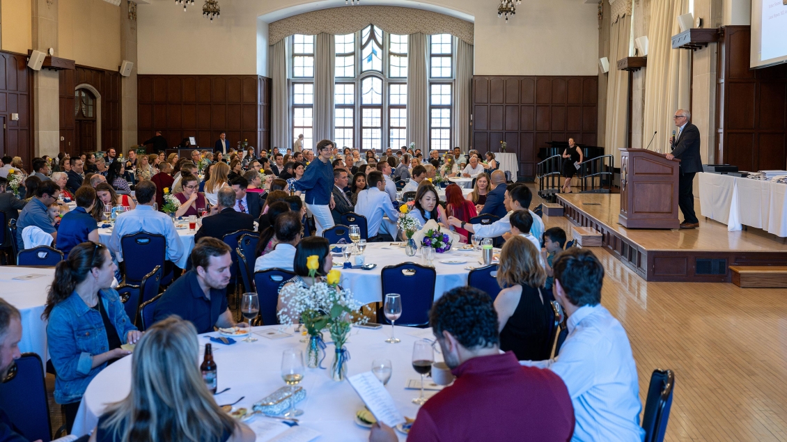 A large group of individuals seated at tables in a spacious room, engaged in conversation and activities.