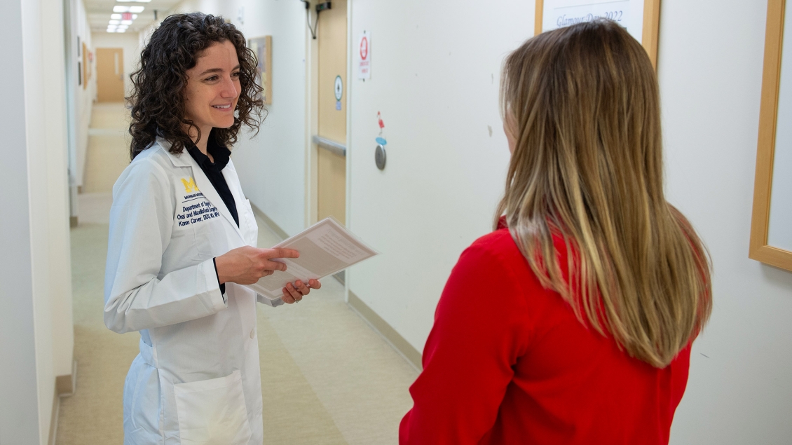 A healthcare professional in a white lab coat, holding some papers, smiles as she speaks with another woman in a red top